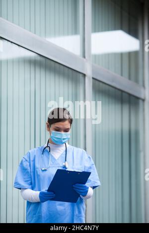 Femme médecin tenant le formulaire de presse-papiers du patient, analysant les données dans le couloir de l'hôpital, portant des gommages PPE de protection bleus:stéthoscope, prote chirurgical Banque D'Images