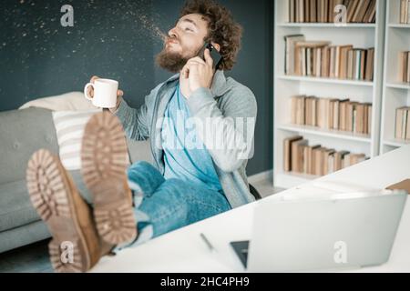 Surpris Man entend les nouvelles sur son smartphone et Spits Out Coffee.L'homme a des jambes sur la table et une tasse de café dans sa main.Gros plan.Arrière-plan des étagères.Photo de haute qualité Banque D'Images