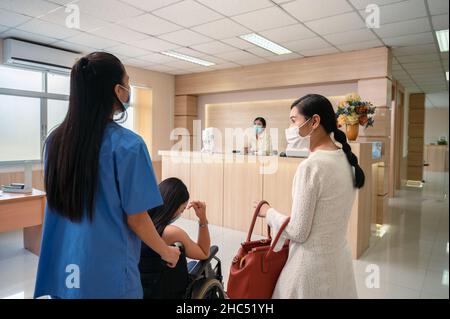 Service d'infirmière poussant la patiente asiatique en fauteuil roulant à contacter la réceptionniste pour qu'elle soit admise à l'hôpital Banque D'Images