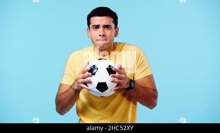 inquiet jeune homme dans un t-shirt jaune tenant le football isolé sur bleu Banque D'Images