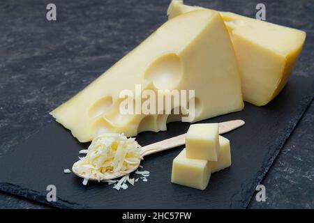 Des quartiers de fromage hollandais et une pile de fromage râpé sur une cuillère en bois sur du noir. Banque D'Images