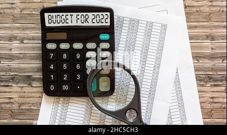 Le mot budget pour 2022 est écrit sur l'écran d'une calculatrice, les documents de paiement se trouvent sur un fond en bois.Accumuler de l'argent et planifier un bu Banque D'Images