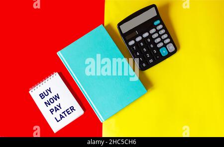 Bloc-notes avec les mots ACHETER MAINTENANT PAYER PLUS TARD sur un fond rouge et jaune avec un journal vert et une calculatrice Banque D'Images