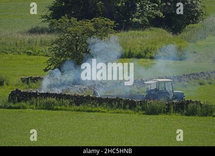 Paysage rural d'été avec des fumées de feu de camp en hausse dans le coin du champ avec agriculteur, tracteur et remorque en présence - Cumbria, Angleterre, Royaume-Uni Banque D'Images