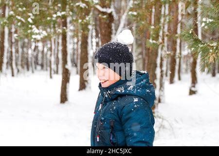 Reflous l'enfant souriant dans la forêt d'hiver en pleine tempête de neige avec boule de neige sur la tête.Drôle de gamin sur fond enneigé d'hiver.Passer activement du temps à l'extérieur. Banque D'Images