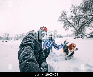 Couple heureux avec leur chien beagle, prenant un selfie pendant une tempête de neige, équipé de vêtements d'hiver, de gros plan, concept de vacances Banque D'Images