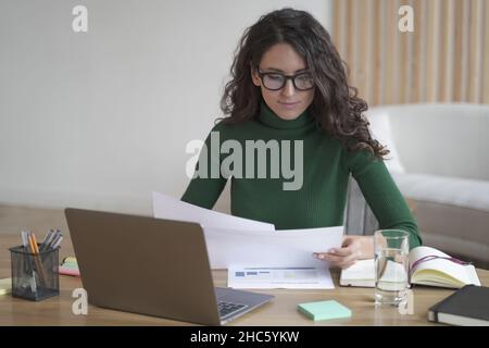 Femme italienne indépendante réfléchie en lunettes, assise au bureau, tenant des papiers et préparant un rapport Banque D'Images