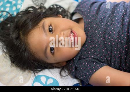 Une belle petite fille indonésienne souriante avec une rangée de dents blanches et de longs cheveux noirs sur le lit, tirer à partir d'un angle élevé Banque D'Images