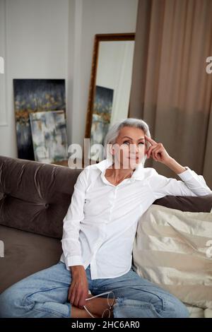 Grand angle de charmante femme d'âge moyen avec cheveux gris et jeans et chemise blanche assis avec des jambes croisées sur le canapé à la maison et souriant à l'appareil photo Banque D'Images