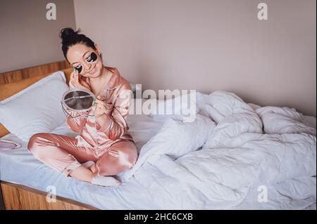 Cosmétologie, soins de la peau, soin du visage, spa, concept de beauté naturelle.Souriante bonne jeune femme en pyjama de soie rose est assise sur le lit avec miroir appliquant des taches hydratantes noires sur les yeux.Routine beauté Banque D'Images
