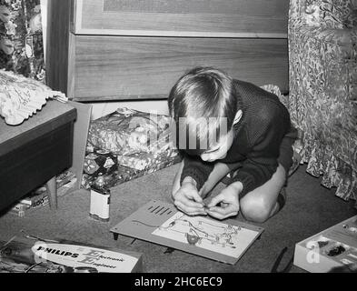 1960s, historique, à l'intérieur d'une pièce, un jeune garçon s'agenouillant sur le sol en jouant avec un cadeau de noël, une carte de circuit imprimé d'un kit d'ingénieur électronique Philips, Angleterre, Royaume-Uni.Les kits de « jeunes ingénieurs » en boîte étaient une activité éducative et ludique populaire que les jeunes garçons devaient entreprendre à cette époque. Banque D'Images