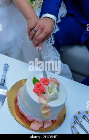 mariée et marié couper le gâteau de mariage.Le gâteau est décoré de roses beige et pêche. Le marié est habillé en costume de mariage bleu et mariée dans un W Banque D'Images