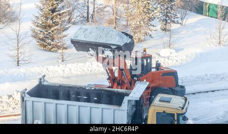 Un gros tracteur orange nettoie la neige de la route et la charge dans le camion.Nettoyage et nettoyage des routes de la ville de la neige en hiver Banque D'Images