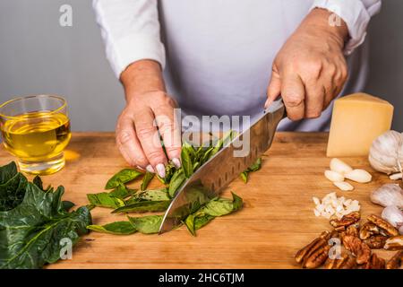 Vue rognée d'une femme adulte méconnue qui hache du basilic avec un couteau de cuisine sur une table en bois pour la préparation d'une sauce au pesto. Banque D'Images
