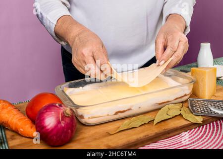 Mains d'une femme méconnaissable qui fait de la lasagne dans un plateau en verre avec des ingrédients crus autour. Banque D'Images