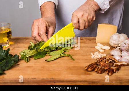Vue rognée d'une femme adulte méconnue coupant du basilic avec un couteau en plastique sur une table en bois pour la préparation d'une sauce au pesto. Banque D'Images