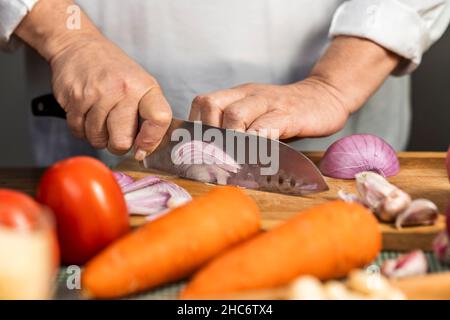 Vue rapprochée des mains d'une femme adulte coupant un oignon avec un couteau de cuisine sur une planche en bois pour la préparation de lasagnes. Banque D'Images