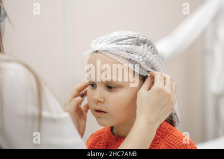 gros plan des mains d'esthéticienne mettant blanc capuchon médical jetable sur la tête de six ans-fille dans le bureau de cosmétologie, se préparant pour le perçage d'oreilles Banque D'Images