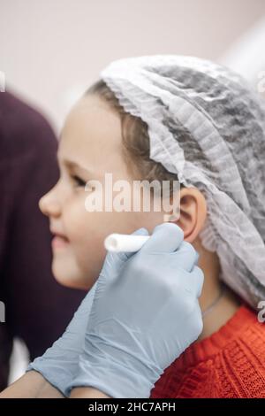 Processus de perçage des oreilles.Le cosmétologue en gants jetables tire un point sur l'oreille au niveau du site de ponction avec un marqueur Banque D'Images