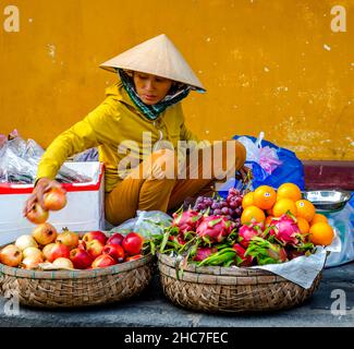 Fruit Market dame portant un dessus de couleur moutarde correspond au mur arrière où elle est assise sur un trottoir à Hoi an, Vietnam Cental Market. Banque D'Images