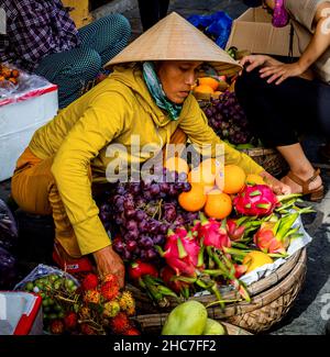 Une dame vietnamienne portant un chapeau conique et un haut de couleur moutarde ramasse son panier de fruits qui est très lourd et surchargé de la terre. Banque D'Images