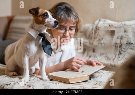 Une femme caucasienne âgée est allongée sur un canapé avec un chien intelligent Jack russell terrier portant des lunettes et une cravate et lisant un livre. Banque D'Images