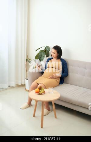 Bonne fille enceinte utilisant la tablette, la navigation sur Internet, manger des fruits à la maison Banque D'Images