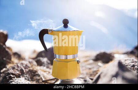 Café en moka jaune avec vapeur au camping de montagne de neige.Pique-nique le matin, personne cuisant une boisson chaude pour le petit déjeuner dans la nature Banque D'Images