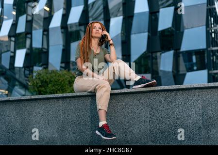 Une jeune fille aux cheveux rouges s'assoit sur un parapet sur le fond d'un immeuble de bureau inhabituel et parle au téléphone avec un collègue.Extérieur Banque D'Images