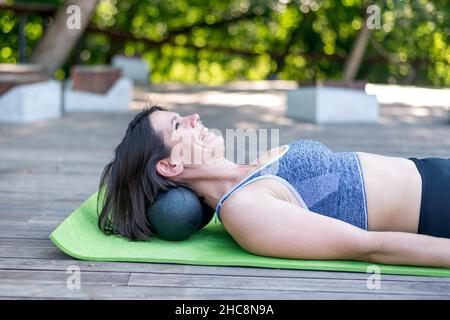 Une jeune fille sportive gaie se trouve sur un tapis de yoga et fait un massage du cou avec des boules myofasciales.Kneads les muscles du cou avec le rouleau MFR à l'extérieur. Banque D'Images