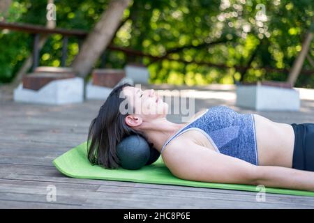 Une jeune fille athlétique se trouve sur un tapis de yoga et fait un massage du cou avec des boules myofasciales.Kneads les muscles du cou avec le rouleau MFR à l'extérieur. Banque D'Images