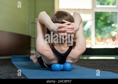 La fille se trouve sur le tapis de yoga et fait un massage de dos avec rouleau pour la physiothérapie.Relâchement myofascial avec boule en mousse.Vue arrière. Banque D'Images