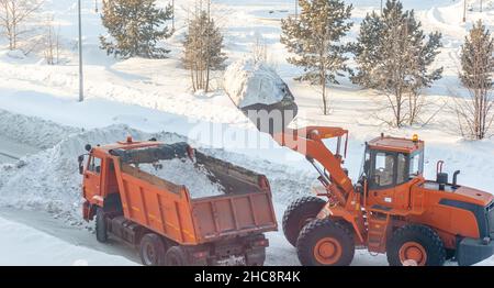 Un gros tracteur orange nettoie la neige de la route et la charge dans le camion.Nettoyage et nettoyage des routes de la ville de la neige en hiver Banque D'Images