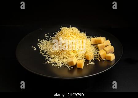 Pile de fromage gouda râpé mince sur une plaque en céramique noire, isolée sur fond noir. Banque D'Images