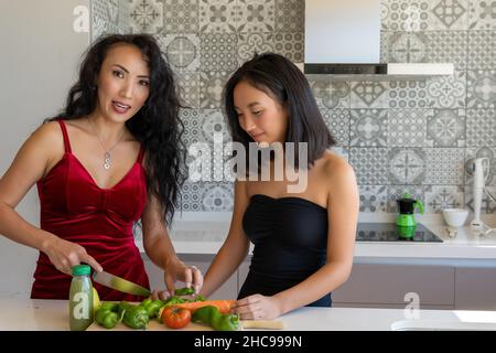 deux femmes cuisinant dans la cuisine portant des robes, l'une d'entre elles coupant des légumes, fond à motifs horizontaux Banque D'Images