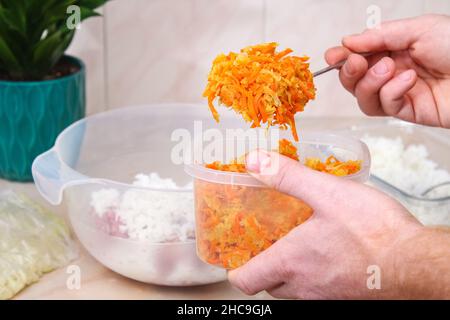 Un homme ajoute des oignons frits et des carottes à la viande hachée dans un bol pour les petits pains au chou paresseux sur la table. Banque D'Images