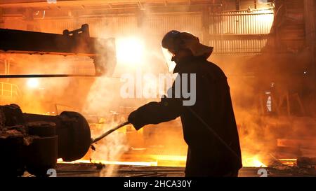 Aciérie découpant les scories du four de fusion de creusets à induction électrique à l'usine métallurgique, atelier de travail dur.Homme dans le masque de protection Banque D'Images