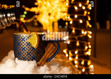 Versez une tasse de thé de thermos avec une tranche de citron et de cannelle dans la neige pendant une nuit d'hiver, avec des arbres illuminés flous en arrière-plan. Banque D'Images