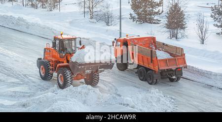 Un gros tracteur orange nettoie la neige de la route et la charge dans le camion.Nettoyage et nettoyage des routes de la ville de la neige en hiver Banque D'Images