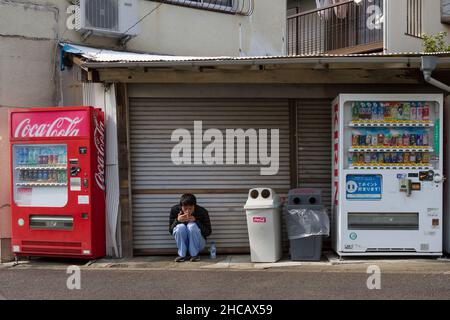 Un jeune Japonais s'accrouille entre les distributeurs automatiques de la salle de Machida, Tokyo, Japon. Banque D'Images