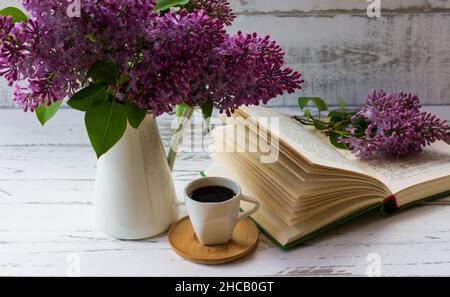 un bouquet de lilas de jardin dans une carafe à côté d'un livre ouvert et d'une tasse de café sur fond blanc de planches de bois Banque D'Images