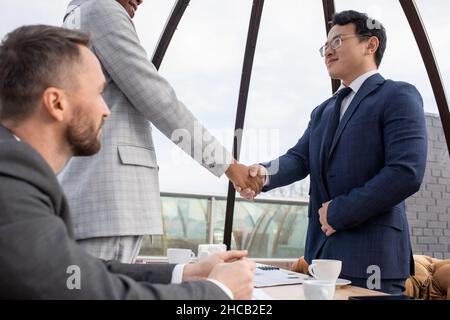 Jeunes partenaires d'affaires contemporains en tenue de cérémonie poignée de main après avoir négocié tout en se tenant debout à table dans un café Banque D'Images