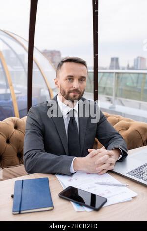 Jeune homme d'affaires contemporain en costume élégant assis près d'une table dans un café et regardant l'appareil photo en attendant un collègue Banque D'Images