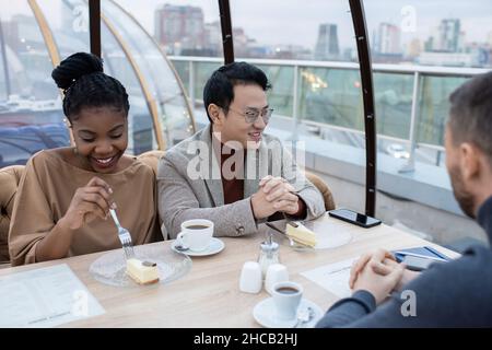 De jeunes collègues interculturels heureux en tenue de formalwear ayant des boissons et des desserts tout en étant assis à table dans le café Banque D'Images