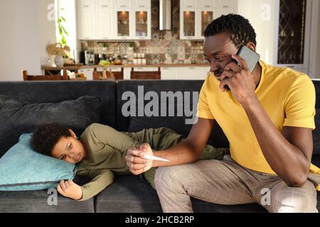 Jeune homme africain avec smartphone à l'oreille appelant médecin pour son petit fils malade tout en étant assis sur un canapé à côté de lui Banque D'Images