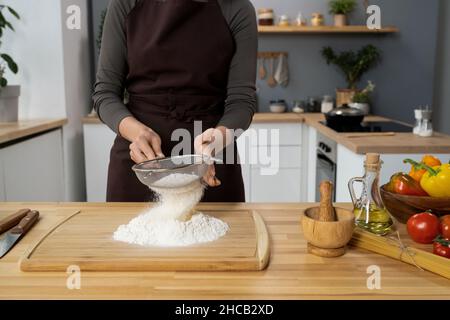 Jeune femme en tablier tamisant la farine à travers le tamis sur le bois tout en préparant la nourriture par table de cuisine Banque D'Images