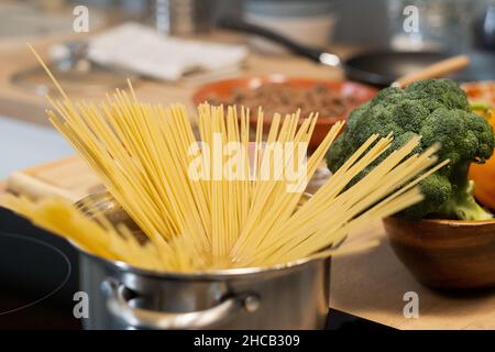 Spaghetti crus dans une casserole métallique sur une cuisinière électrique chaude contre une table de cuisine avec des légumes frais Banque D'Images