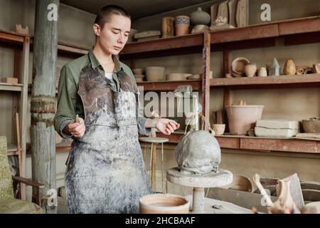 Jeune femme en tablier debout et regardant sa sculpture, elle apprend à la faire de l'argile en studio de céramique Banque D'Images