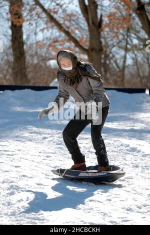 Une femme active dans ses 50s traîneaux sur une colline enneigée pendant un hiver froid.Dans un parc à Flushing, Queens, New York. Banque D'Images