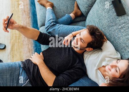 Un jeune couple heureux prend le selfie à travers un smartphone sur un canapé à la maison Banque D'Images
